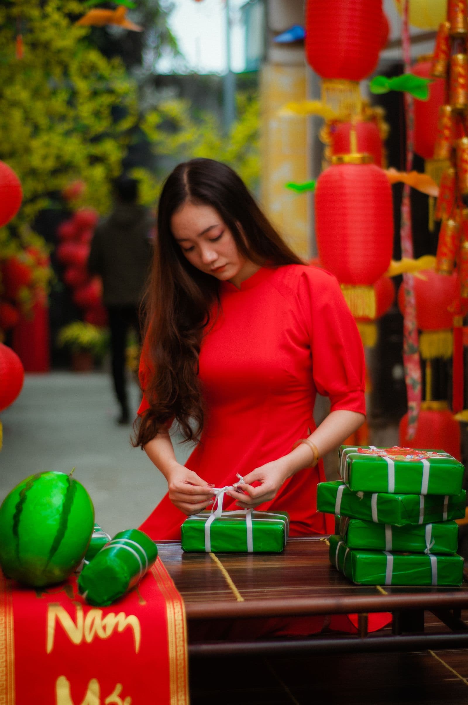 Người mặc áo dài đỏ buộc dây những hộp quà xanh ngày Tết, đèn lồng phía sau