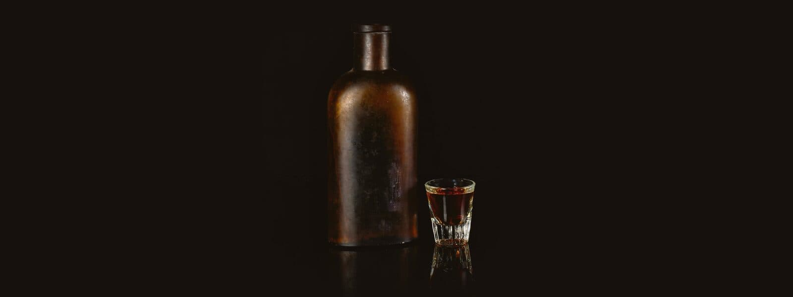 Chai thuỷ tinh cũ không nhãn và một ly nhỏ rượu màu hổ phách trên nền đen